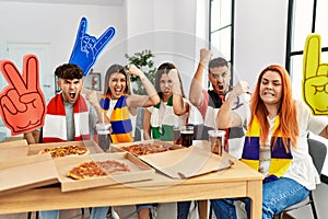 Group of young hispanic people eating pizza supporting soccer team at home annoyed and frustrated shouting with anger, yelling