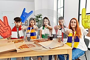 Group of young hispanic people eating pizza supporting soccer team at home pointing with finger to the camera and to you,