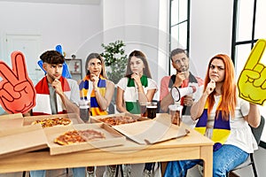 Group of young hispanic people eating pizza supporting soccer team at home serious face thinking about question with hand on chin,