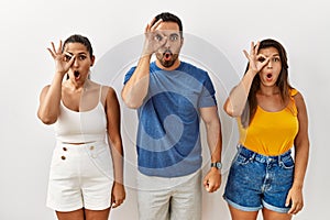 Group of young hispanic people standing over isolated background doing ok gesture shocked with surprised face, eye looking through