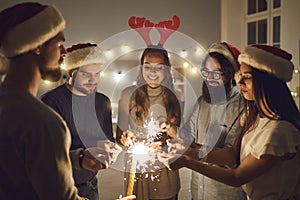 Group of young male and female friends stand in a circle and light sparklers on New Year`s Eve.