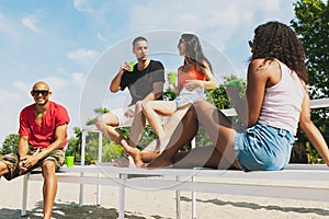 Group of young people, men and women resting on the beach, drinking cool drinks, having fun sunbathing