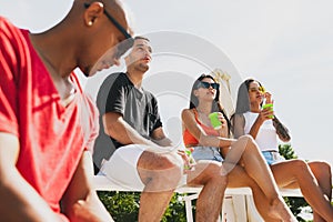 Group of young people, men and women resting on the beach, drinking cool drinks, having fun, sunbathing