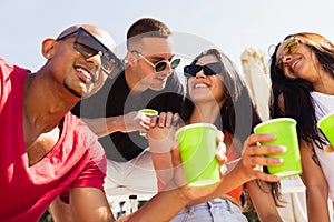 Group of young people, men and women resting on the beach, drinking cool drinks, having fun sunbathing