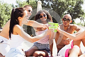 Group of young people, men and women resting on the beach, drinking cool drinks, having fun sunbathing