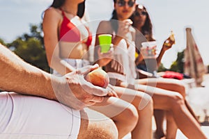 Group of young people, men and women resting on the beach, drinking cool drinks, having fun, sunbathing