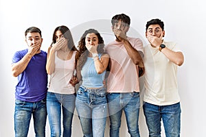Group of young people standing together over isolated background shocked covering mouth with hands for mistake