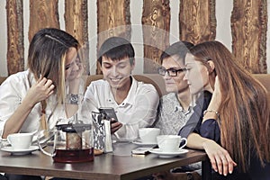 Group of young people from two couples of guys and girls have a tea time in the cafe and looking on the smartphone absorbedly