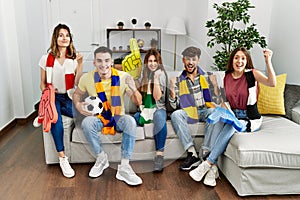 Group of young people watching football game wearing team scarf cheering game sitting on the sofa screaming proud, celebrating