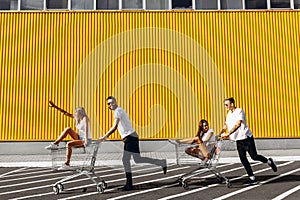 A group of young people in white t-shirts, fun ride on carts near the store, supermarket, friends have fun in Sunny weather