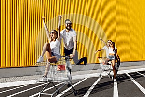 A group of young people in white t-shirts, fun ride on carts near the store, supermarket, friends have fun in Sunny weather