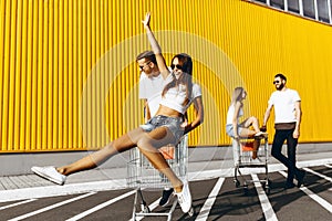 A group of young people in white t-shirts, fun ride on carts near the store, supermarket, friends have fun in Sunny weather