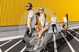 A group of young people in white t-shirts, fun ride on carts near the store, supermarket, friends have fun in Sunny weather