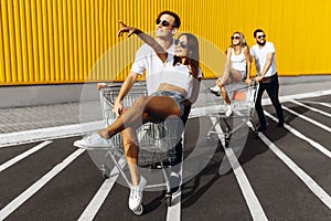 A group of young people in white t-shirts, fun ride on carts near the store, supermarket, friends have fun in Sunny weather