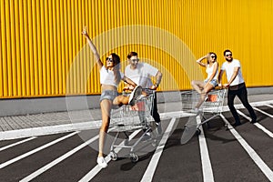 A group of young people in white t-shirts, fun ride on carts near the store, supermarket, friends have fun in Sunny weather