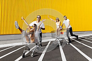 A group of young people in white t-shirts, fun ride on carts near the store, supermarket, friends have fun in Sunny weather