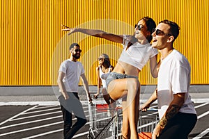 A group of young people in white t-shirts, fun ride on carts near the store, supermarket, friends have fun in Sunny weather