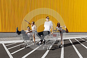 A group of young people in white t-shirts, fun ride on carts near the store, supermarket, friends have fun in Sunny weather