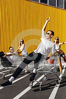 A group of young people in white t-shirts, fun ride on carts near the store, supermarket, friends have fun in Sunny weather