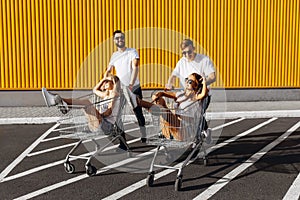 A group of young people in white t-shirts, fun ride on carts near the store, supermarket, friends have fun in Sunny weather