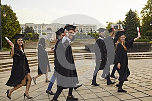 Group of young smiling university graduates in traditional mantles walking, holding diplomas in raised hands