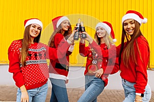 Group of young women in Santa Claus hats, friends cheerfully celebrate New Year and drink drinks in glass bottles, on the