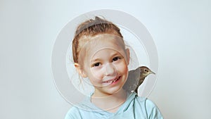 Grown up starling nestling sit on shoulder of little girl and sing with open beak. Portrait of happy child on gray.