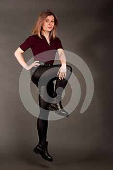 Growth Portrait of the blonde in the red sweater with short sleeves, leather trousers and boots. Studio shot on dark gray