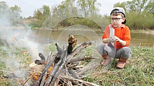 The grubby little kid is squatting near a burning fire on the background of a river in the spring