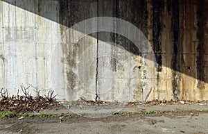 Grunge concrete block wall with moss and dirty divided in two by shadow. Road side with weeds and waste in front.
