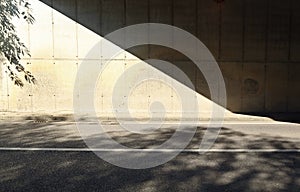 Grunge concrete wall of an underpass divided in two by the shadow of the bridge. Sidewalk and two lane road in front.