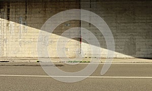 Grunge gray concrete wall of an underpass divided in two by the shadow of the bridge. Cement sidewalk and two lane road in front.