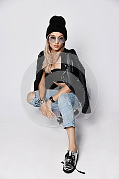 Grunge style woman in torn jeans, bralet, silver boots with untied shoelaces and knitted hat sitting squatting on white