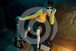 Grunge style young hipster woman with curly hair posing with red guitar and amp in neon lights. 90s style concept.