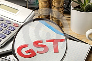 GST word in notebook under magnifying glass and calculator, stack of coins on brown wooden background