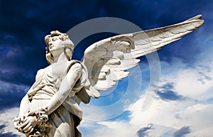 Guardian angel white marble sculpture with open long wings across frame against bright sunny dark blue sky with white clouds.