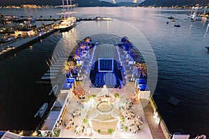 Guests sit at tables on the pier near a festively lit stage in front of a long seaside pool. Drone