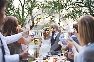 Guests with smartphones taking photo of bride and groom at wedding reception outside.