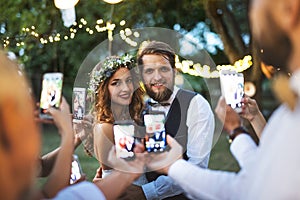 Guests with smartphones taking photo of bride and groom at wedding reception outside.