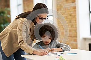 Guiding young female teacher in glasses helping little boy with the task. Kid studying in elementary school, sitting at