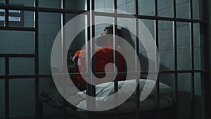Guilty criminal in orange uniform sits on the bed in prison cell