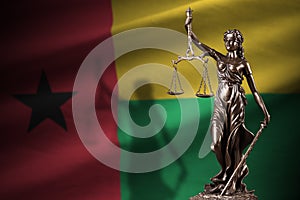 Guinea Bissau flag with statue of lady justice and judicial scales in dark room. Concept of judgement and punishment