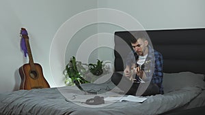 Guitar player playing on guitar at home on bed. Musician in headphones plays on electric guitar. Young male millennial is practici
