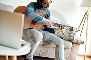 Guitar player. Young man playing guitar while sittting at home and watching video tutorial on laptop online. Distance