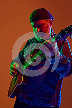 Guitarist with beard intently adjusts electric guitar in colorful neon light against orange studio background.