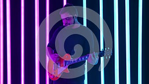 The guitarist enjoys his electric guitar playing in a dark studio surrounded by bright blue and pink neon tubes. A man