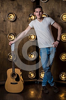 Guitarist, music. A young man stands with an acoustic guitar in the background with the lights behind him. Vertical frame