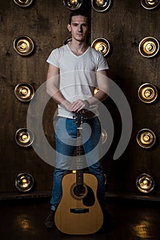 Guitarist, music. A young man stands with an acoustic guitar in the background with the lights behind him. Vertical frame