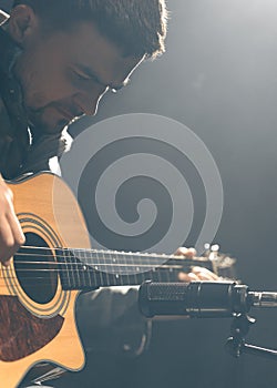 A guitarist plays an acoustic guitar with a microphone on a dark background.