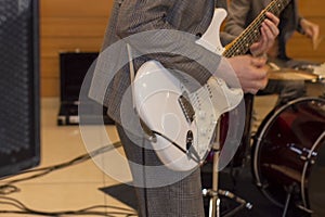 Guitarist plays solo. Grey suit and white guitar. There`s a drum kit in the background